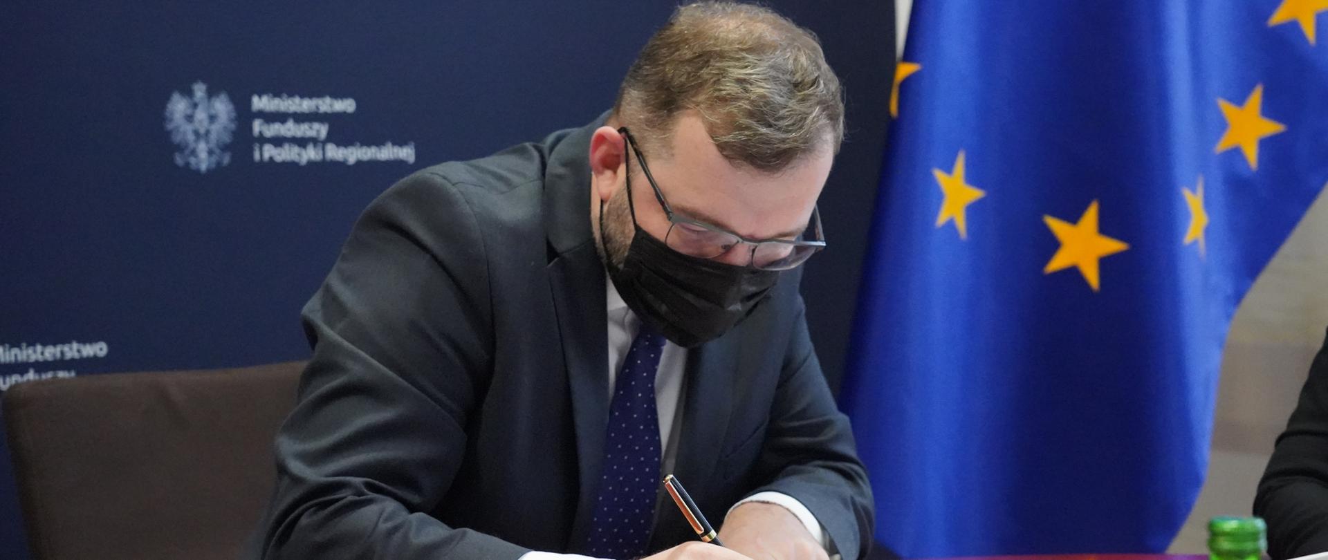 Minister Grzegorz Puda podpisuje dokument, w tle ścianka z logo Ministerstwa funduszy i polityki regionalnej oraz flaga Unii Europejskiej