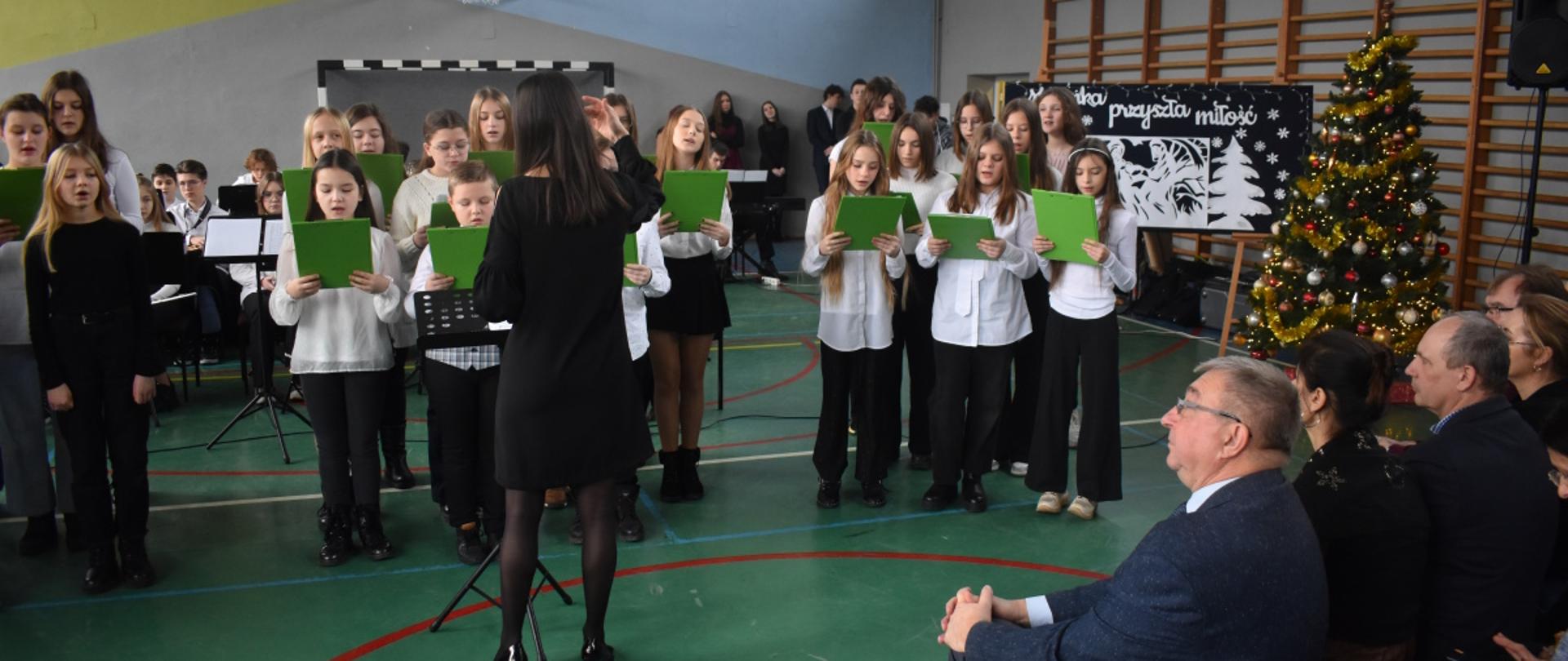 Na zdjęciu, w sali gimnastycznej ZST w Jaśle, widnieje chór szkolny, którym dyryguje Monika Twarduś. Na pierwszym planie widoczne również Grono Pedagogiczne ZST. Kolorystyka zdjęcia: biało-żółto-zielono-niebiesko-czarna. 