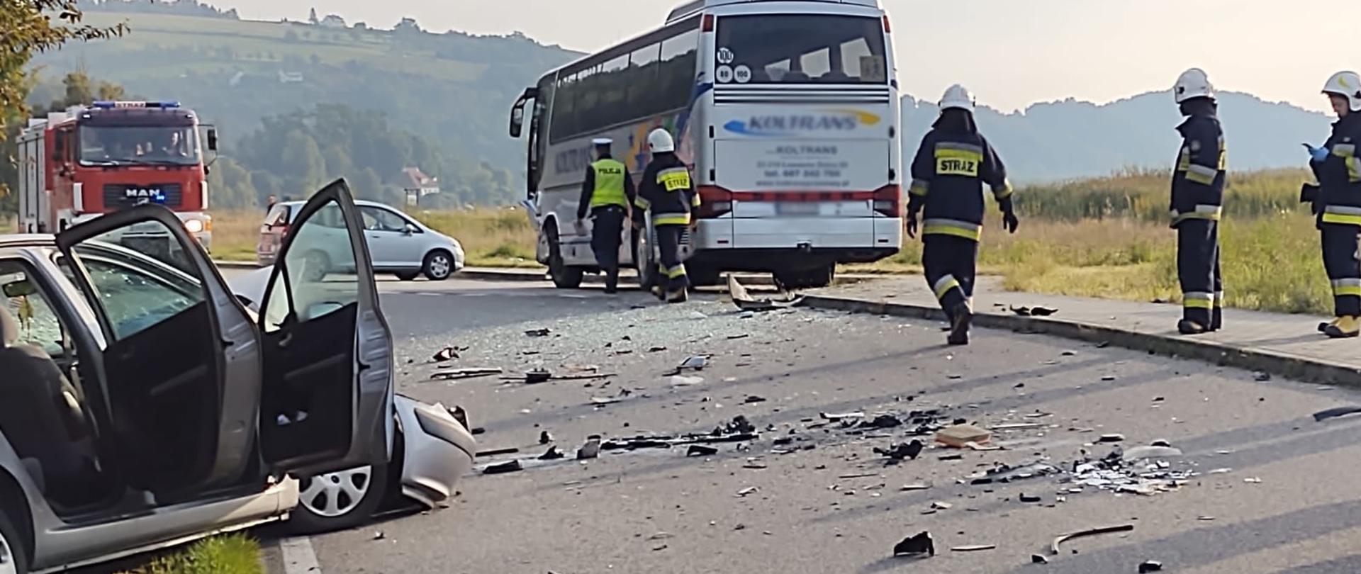 Samochód osobowy rozbity poza drogą. Na drodze rozsypane części samochodu. Na chodniku stoi autobus. Strażacy i Policjanci prowadzą działania. W tle samochód pożarniczy.