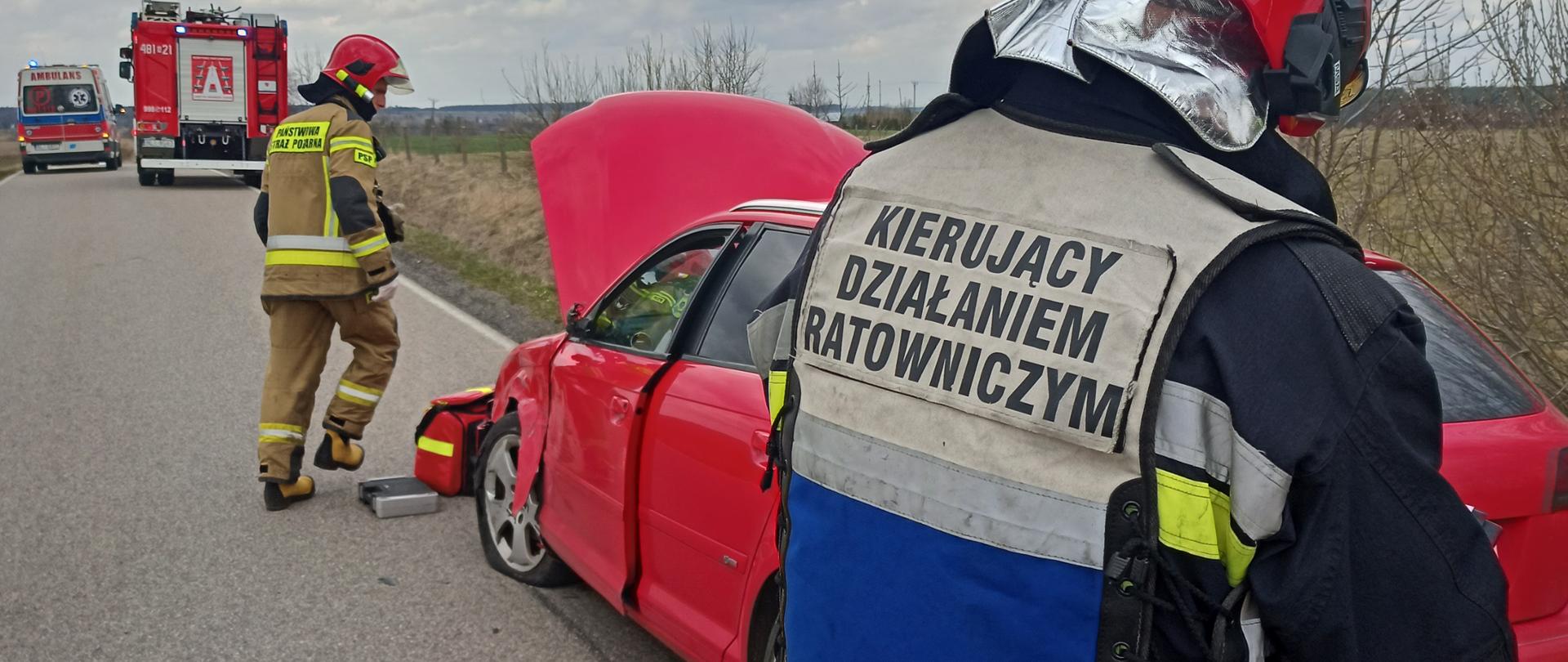 Zdjęcie przedstawia uszkodzony pojazd oraz KDRa i strażaka PSP, który zabezpiecza pojazd. W tle pojazd pożarniczy oraz karetka ZRM