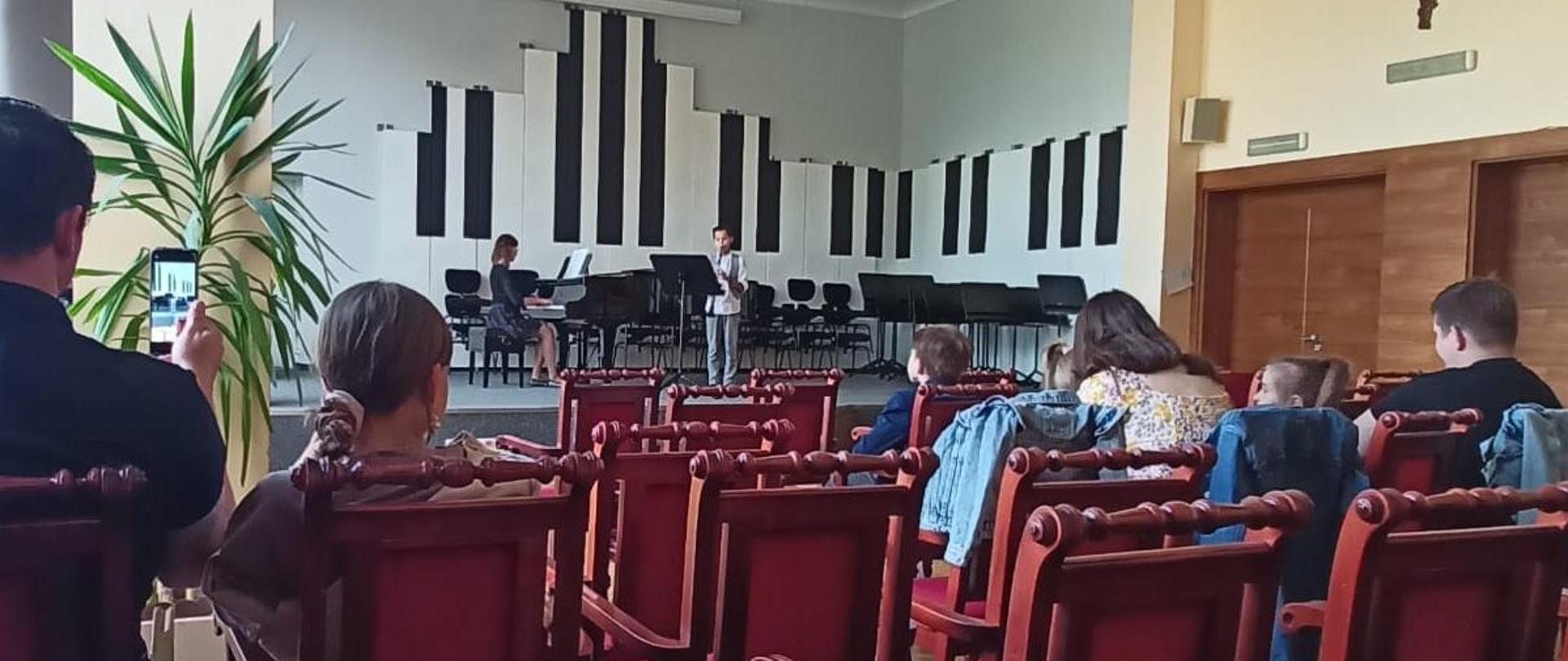 Chłopiec gra na klarnecie, kobieta akompaniuje. Z przodu widoczna od tyłu widownia z publicznością.