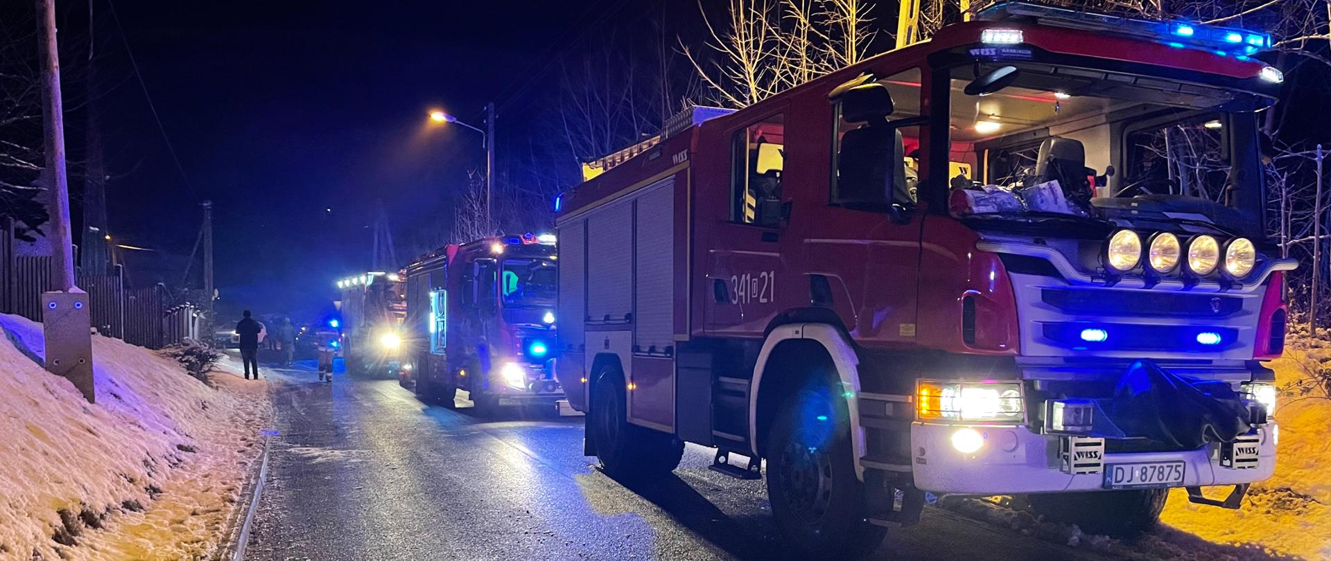 Na zdjęciu widać samochody strażacki z włączonymi światłami ostrzegawczymi. Samochody znajdują się na jezdni. Zdjęcie wykonane w nocy. 