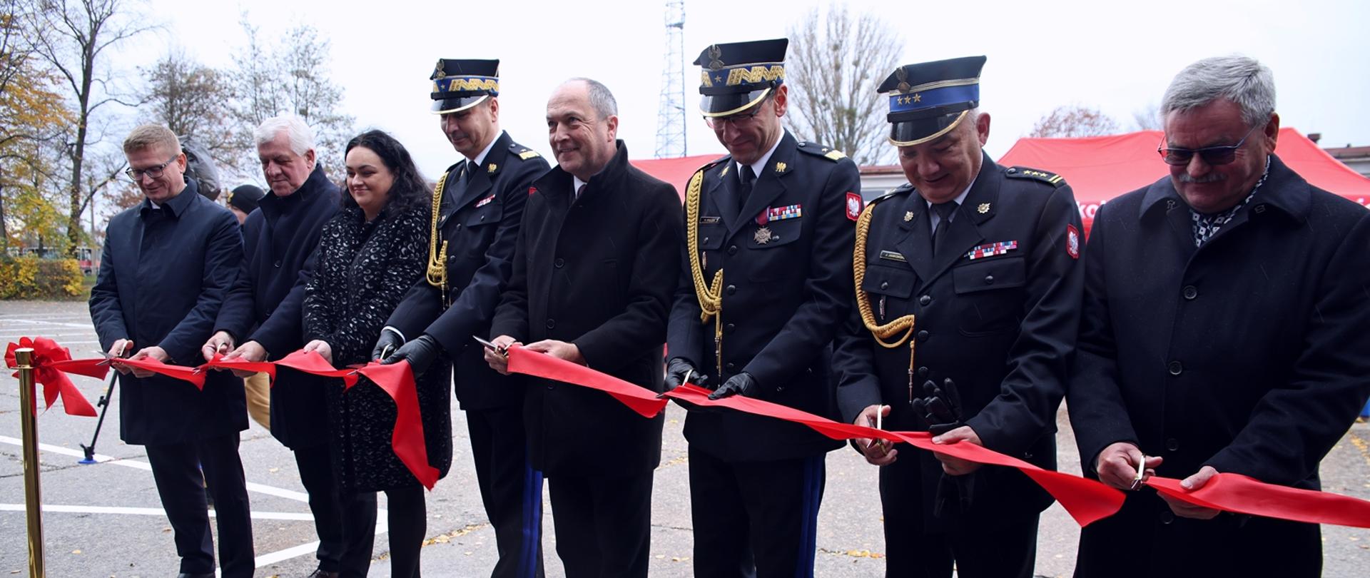 Przecięcie czerwonej wstęgi podczas uroczystości otwarcia nowego obiektu Centralnej Szkoły PSP. W szeregu stoją przedstawiciele władz, kobieta oraz oficerowie PSP w galowych mundurach, wszyscy trzymają nożyczki i przecinają wstęgę.