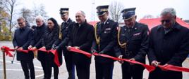Przecięcie czerwonej wstęgi podczas uroczystości otwarcia nowego obiektu Centralnej Szkoły PSP. W szeregu stoją przedstawiciele władz, kobieta oraz oficerowie PSP w galowych mundurach, wszyscy trzymają nożyczki i przecinają wstęgę.