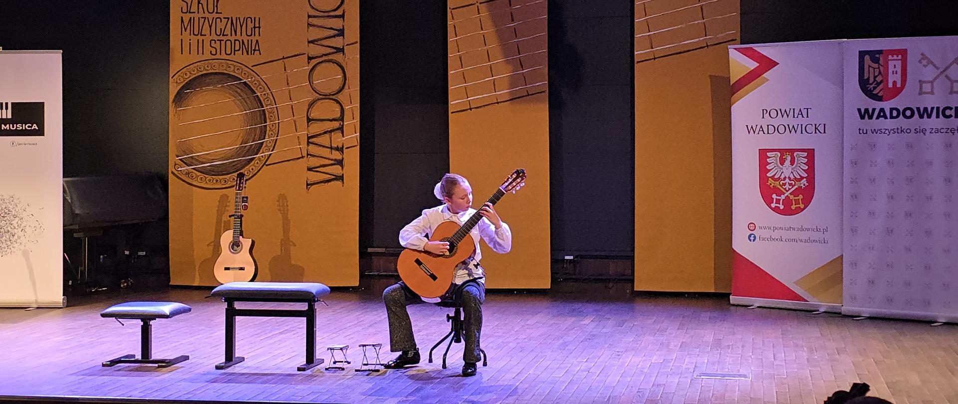 Młoda artystka gra na gitarze klasycznej na dużej scenie podczas występu solowego. Siedzi na profesjonalnym krześle z podnóżkiem, ubrana w białą koszulę i ciemne spodnie. Tło stanowi dekoracja z żółtych pasów materiału z motywami muzycznymi oraz napisami: „V Ogólnopolski Festiwal Gitarowy Szkół Muzycznych I i II Stopnia Wadowice 2026”. Po prawej stronie widoczne są banery promocyjne powiatu wadowickiego i miasta Wadowice.