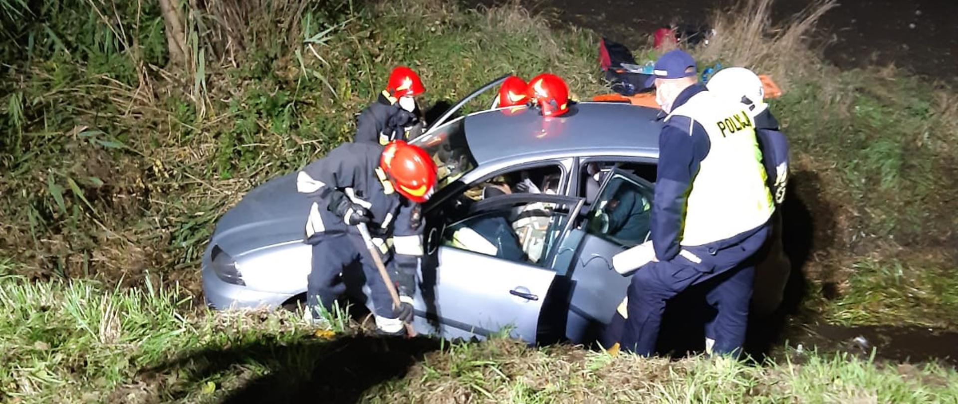 Samochód osobowy marki Seat wjechał do rowu. Przy nim znajduje się trzech strażaków oraz policjant którzy wyciągają poszkodowane osoby. 