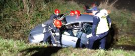 Samochód osobowy marki Seat wjechał do rowu. Przy nim znajduje się trzech strażaków oraz policjant którzy wyciągają poszkodowane osoby. 