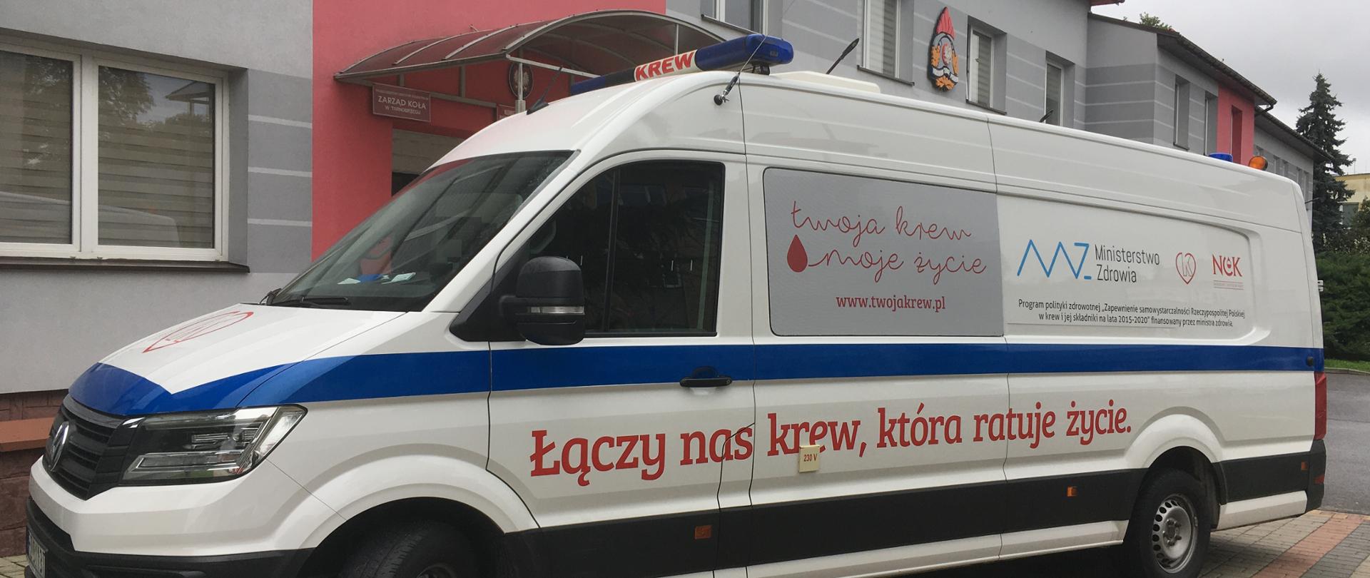 Biały samochód typu bus stoi przed budynkiem. Na samochodzie widoczne napisy związane z oddawaniem krwi.