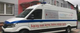 Biały samochód typu bus stoi przed budynkiem. Na samochodzie widoczne napisy związane z oddawaniem krwi.