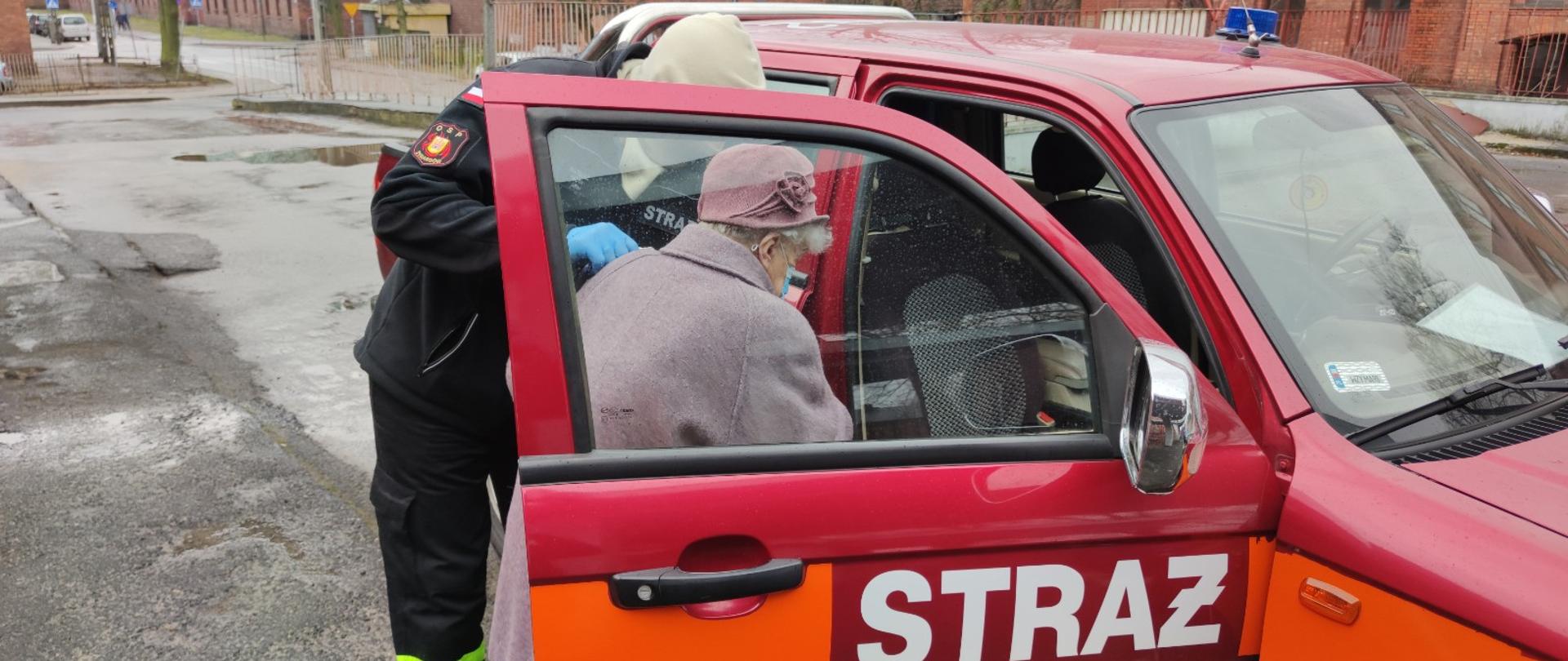 Strażak pomaga starszej kobiecie wsiąść do samochodu strażackiego. W tle budynki z czerwonej cegły.