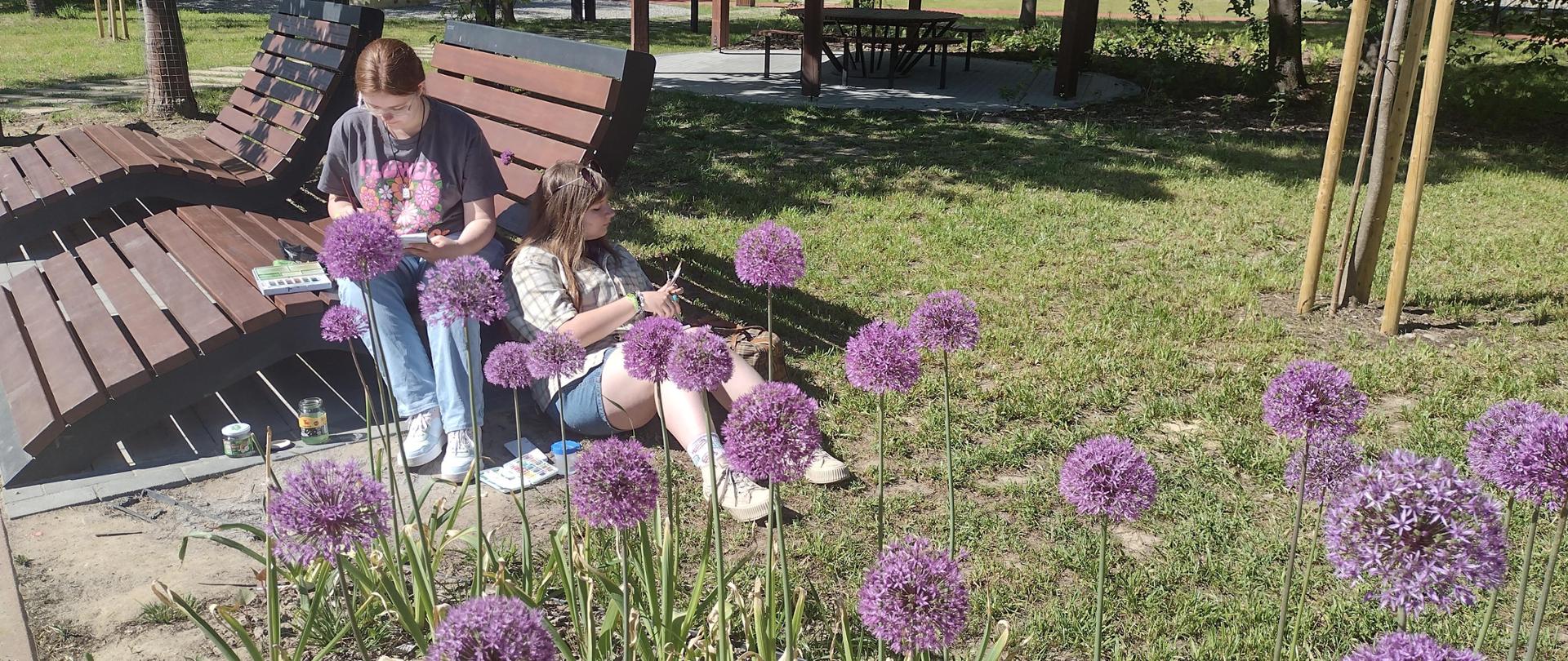 Dwie dziewczyny rysujące w plenerze widziane przez kwiaty