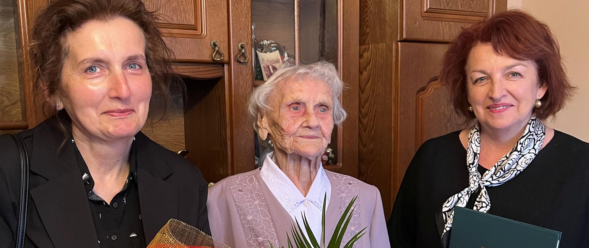 W pokoju na tle regału stoją trzy kobiety – pośrodku Jubilatka Pani Monika Lech, która trzyma w rękach bukiet przepięknych kwiatów . Obok po prawej stronie stoi – Zastępca Kierownika Placówki Terenowej KRUS w Ciechanowie Pani Dorota Tartas, która trzyma w rękach list gratulacyjny Prezesa KRUS. Po lewej stronie stoi Zastępca Kierownika Wydziału Świadczeń Palcówki Terenowej KRUS w Ciechanowie Joanna Ankiewicz-Kołakowska. Wszystkie trzy Panie są pogodne i uśmiechnięte. Starsza Pani jest wzruszona i przejęta całym wydarzeniem. 