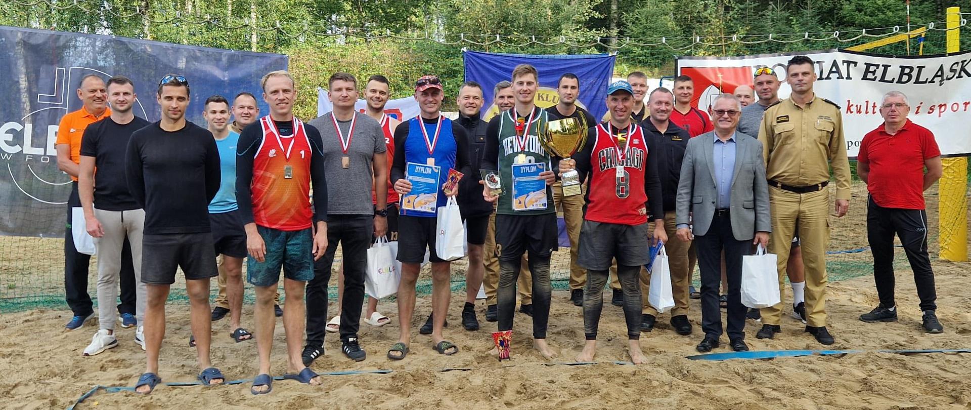 Strażacy stoją w strojach sportowych na piaszczystym boisku do piłki siatkowej. Zwycięzcy turnieju trzymają pamiątkowy puchar. W tle banery reklamowe.