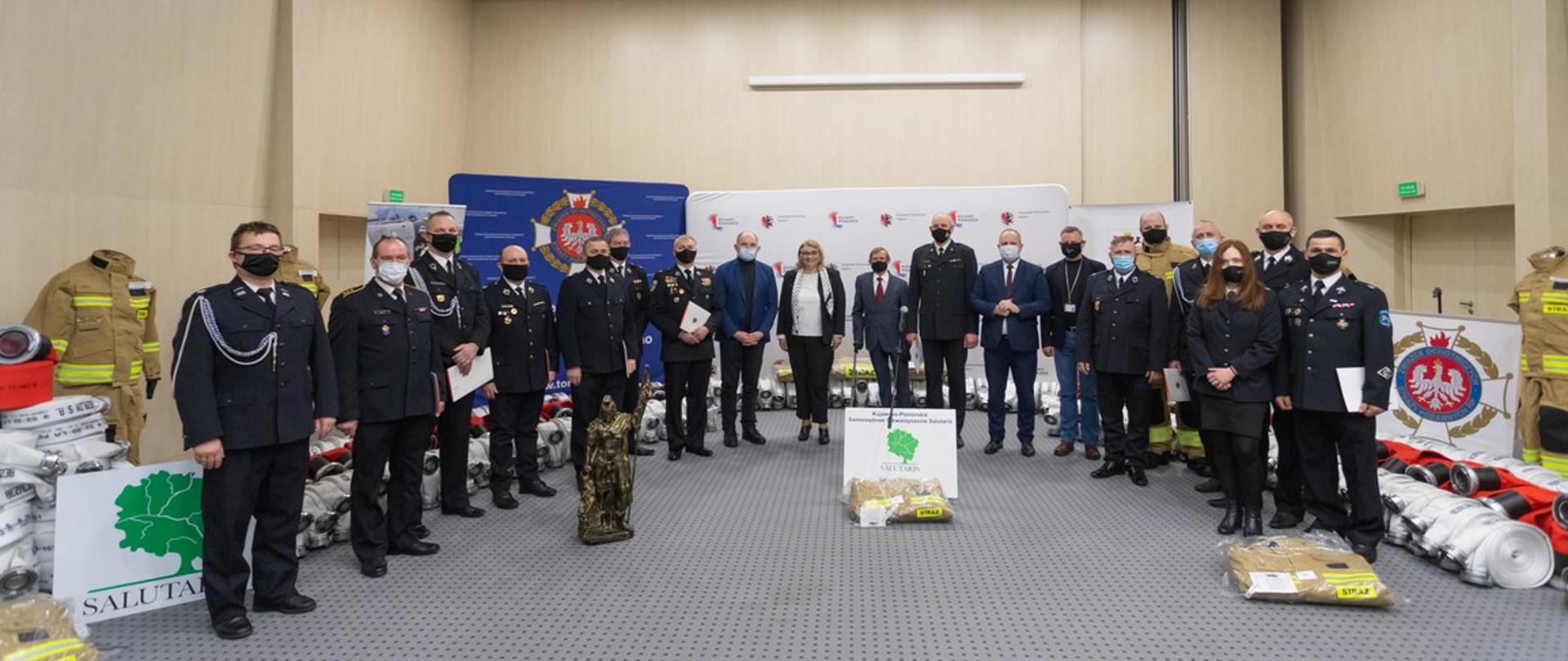 Na zdjęciu w dużym pomieszczeniu ustawieni łukiem strażacy w mundurach OSP jeden w mundurze PSP oraz kilkoro kobiet i mężczyzn w ubraniach cywilnych. W tle stoją ubrania specjalne oraz zwinięte w krąg węże pożarnicze, a także banery.
