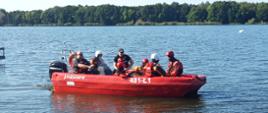 Szkolenia z ratownictwa na obszarach wodnych. Grupa strażaków ubrana w skafandry nurkowe i kamizelki ratunkowe płynie łodzią motorową po jeziorze. W tle jezioro i drzewa.