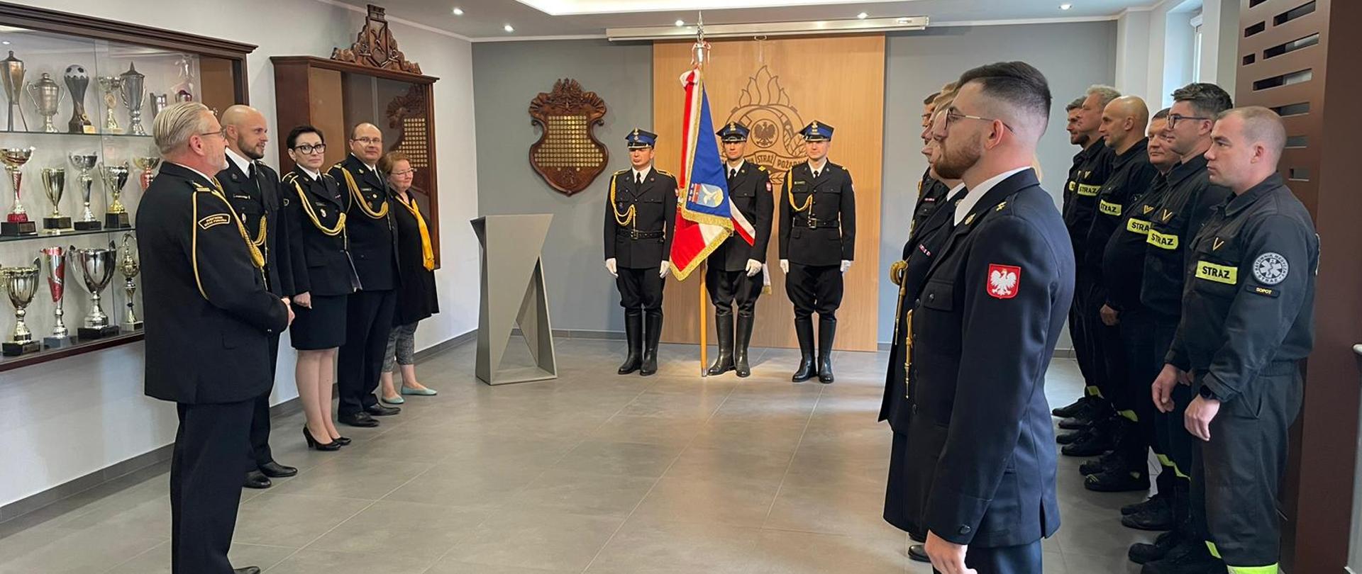 Uroczysta zbiórka ze sztandarem na świetlicy. Komendant wita wszystkich strażaków i pracowników