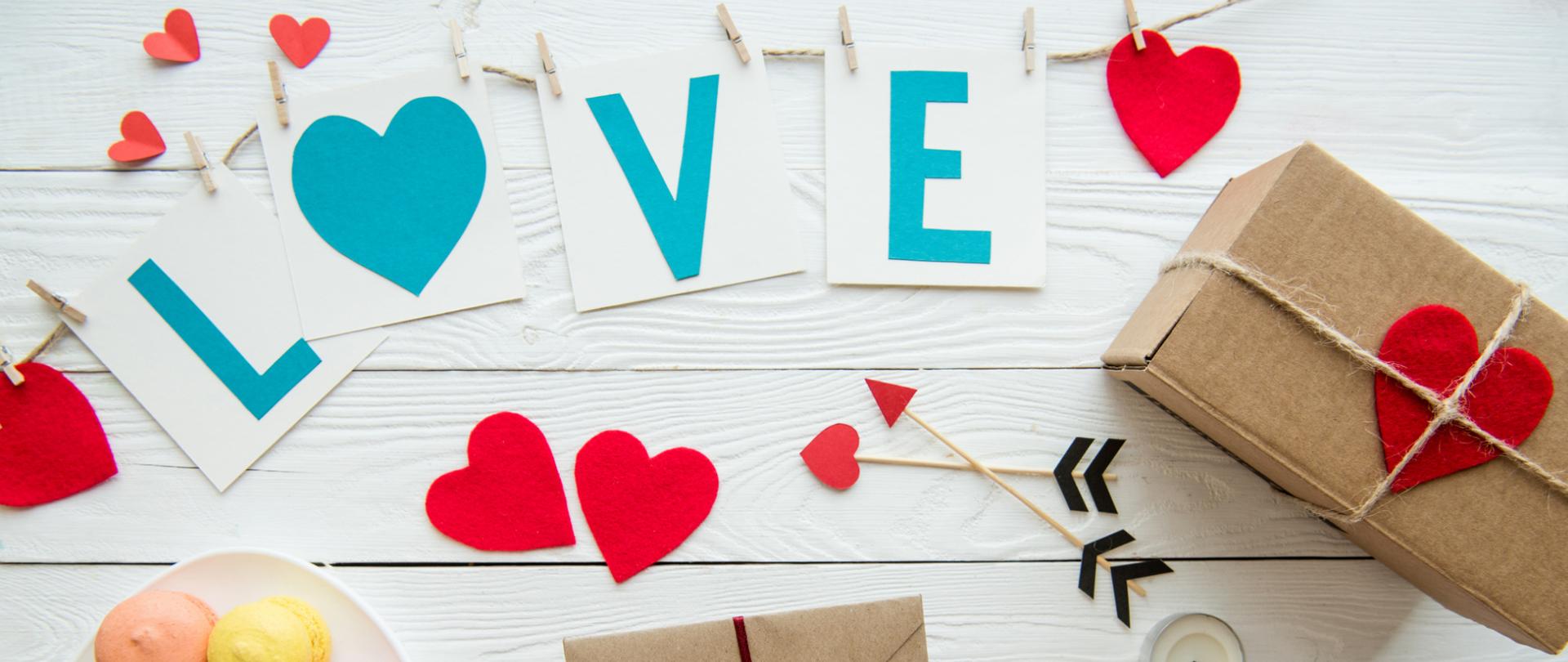 Na zdjęciu jest napis LOVE, wycięte z papieru serduszka, makaroniki znajdujące się na talerzu, prezenty oraz strzały "amora" i świeczka. Wszystko znajduje się na drewnianym blacie.