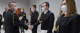 Zastępca komendanta powiatowego w Łowiczu mł. bryg. Rafał Garbarz ubrany w mundur galowy ze sznurem w maseczce ochronnej na twarzy składa gratulacje z okazji awansu na wyższy stopień służbowy strażakowi tutejszej komendy który ubrany jest w mundur galowy ze sznurem w maseczce ochronnej na twarzy, w głębi stoją funkcjonariusze w maseczce ochronnej na twarzy