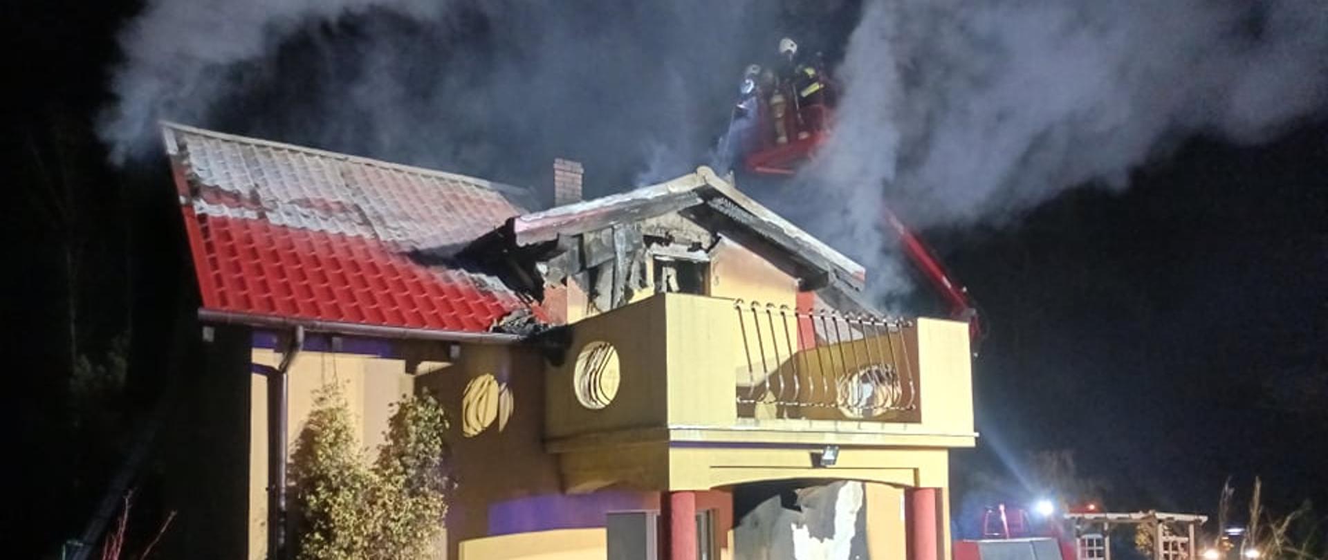 Na zdjęciu widać palący się dach budynku mieszkalnego. Strażacy na podnośniku gaszą pożar.