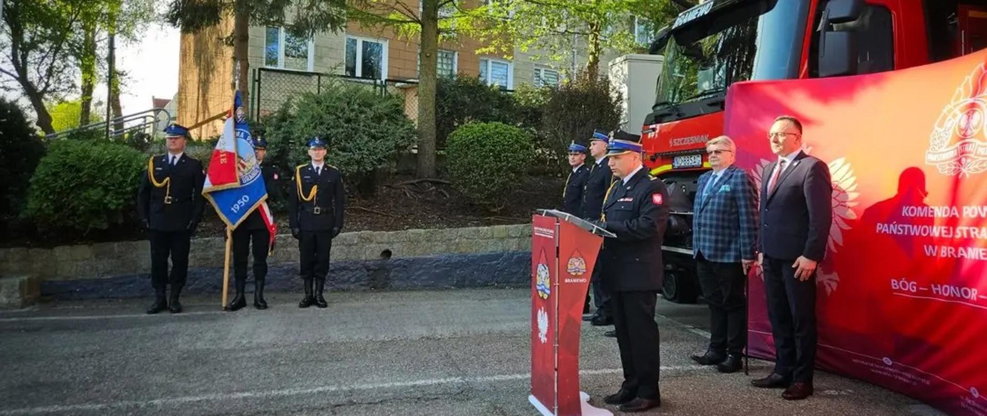 Na wprost na placu stoi sztandar komendy powiatowej PSP w Braniewie. Z prawej strony stoi komendant powiatowy, burmistrz Braniewa oraz starosta braniewski.