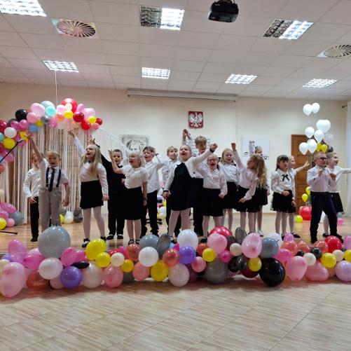 Zdjęcie ukazuje grupę dzieci wykonującą piosenkę na udekorowanej kolorowymi balonami scenie. Dzieci są radosne i uśmiechnięte. Ubrane są w białe bluzki i ciemne spódnice/spodnie. Za dziećmi widoczne jest godło Polski