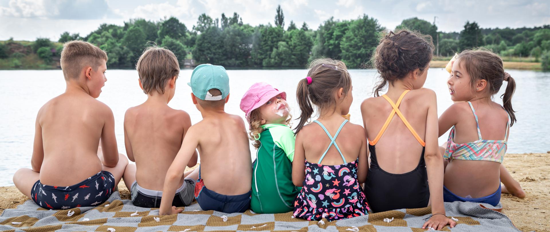 Back view children sit near the river and relax after swimming, sunbathe, eat ice cream.