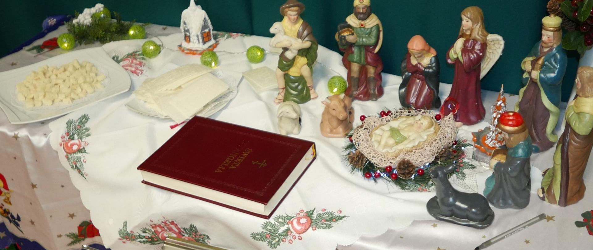 Na stole po prawej stronie ustawione są porcelanowe figurki przedstawiające dzieciątko w żłóbku na środku stołu leży księga święta ewangelia. Po lewej stronie leży na talerzykach opłatek i prosfora.