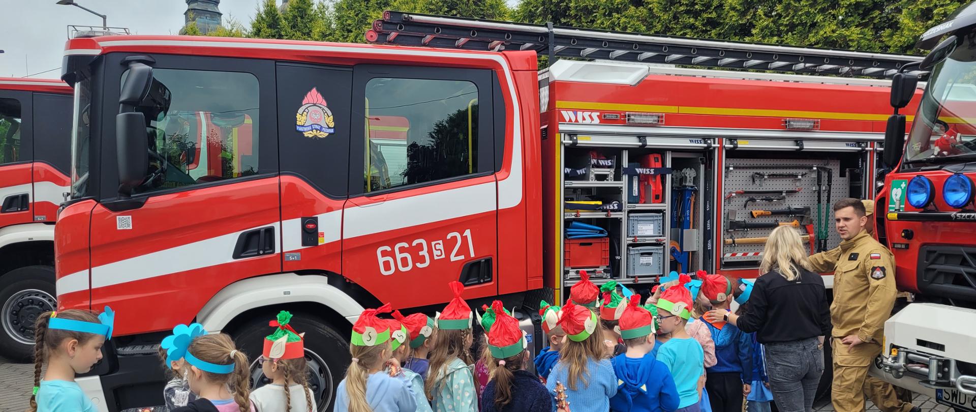 Strażak prezentuje grupie dzieci wyposażenie samochodu ratowniczo-gaśniczego