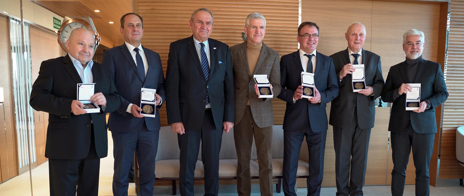 Do zdjęcia pozują elegancko ubrani mężczyźni. Sześciu z nich trzyma w rękach medale 100-lecia Polskiego Komitetu Olimpijskiego. 