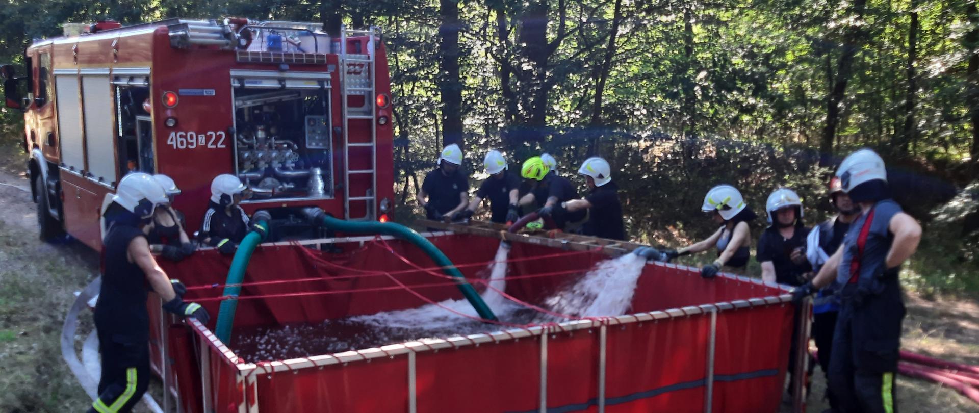 Zdjęcie przedstawia strażaków napełniających zbiornik w czasie ćwiczeń w lesie.