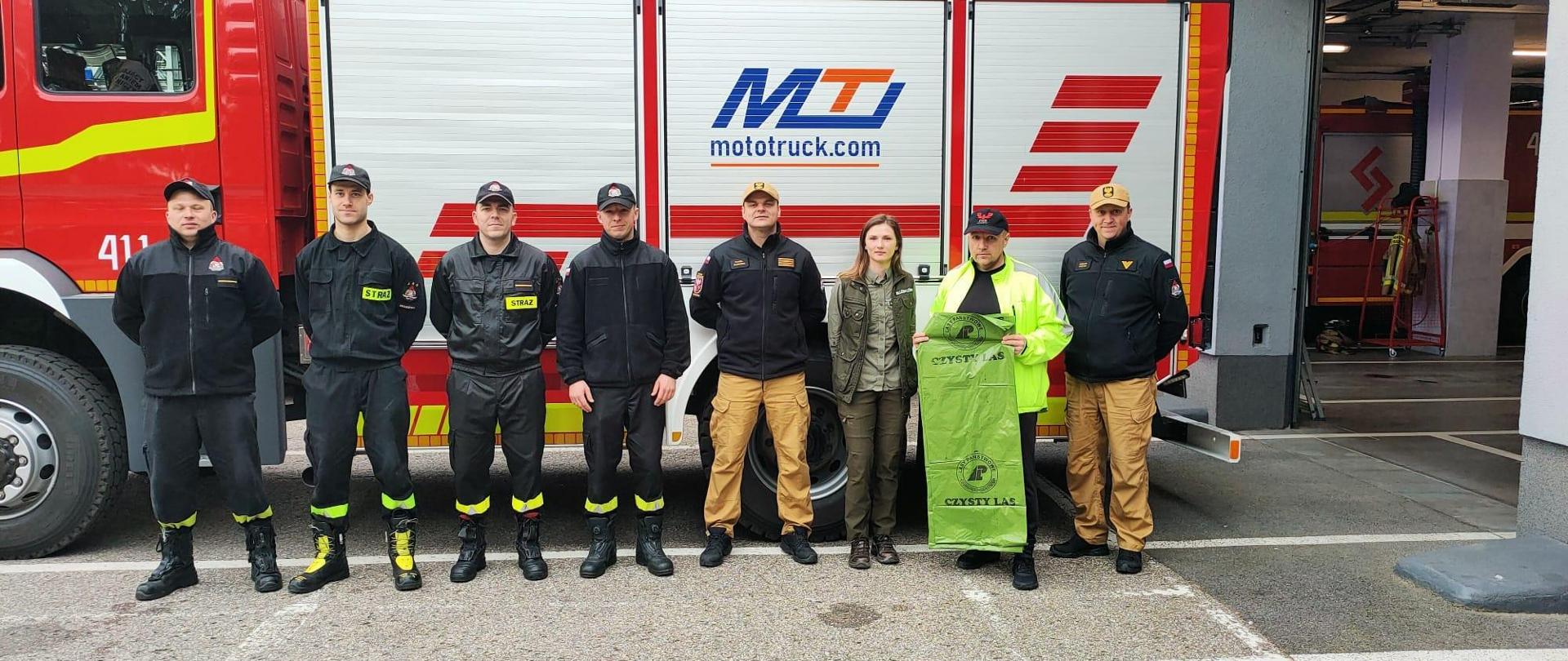 Strażacy ustawieni obok siebie na tle samochodu strażackiego.