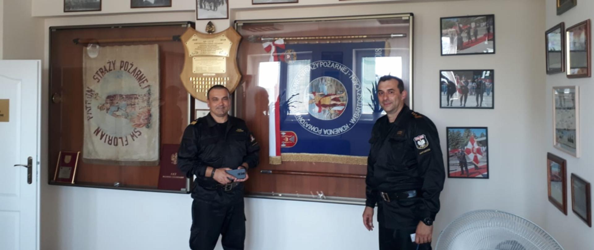 zdjęcie W-M Komendanta Wojewódzkiego i Komendanta Powiatowego PSP w Bartoszycach na tle gabloty ze sztandarami
