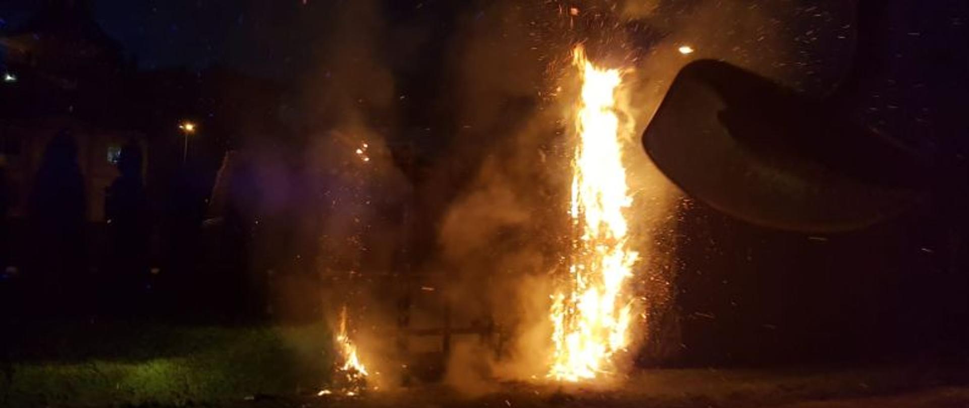 Zdjęcie wykonane z wnętrza samochodu gaśniczego. Widok z kabiny na palące się tuje wzdłuż ogrodzenia boiska Orlik. Tuje są bardzo wysokie, całkowicie objęte ogniem. Część drzew jest spalona. Zdjęcie wykonane po zmierzchu oświetlone przez palące się drzewa