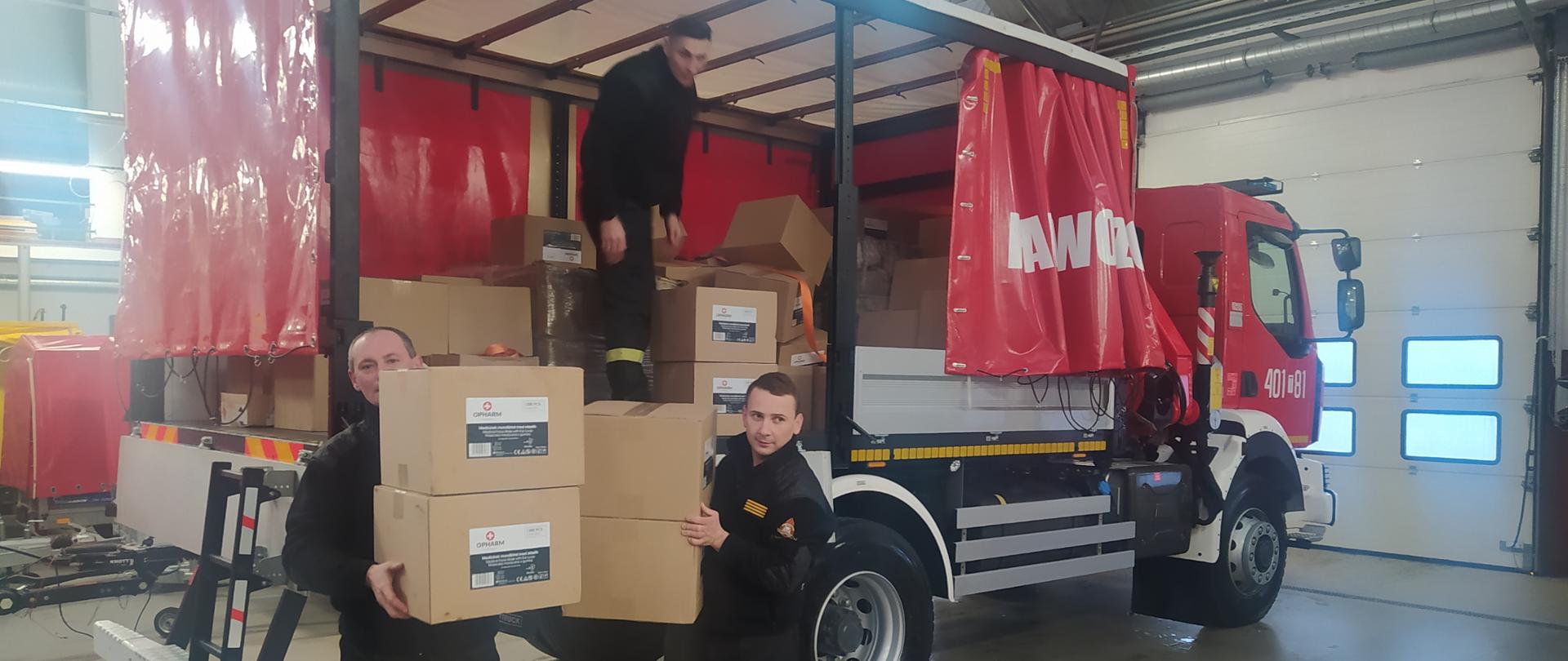 Zdjęcie przedstawia trzech strażaków zdejmujących pudła z samochodu kwatermistrzowskiego 