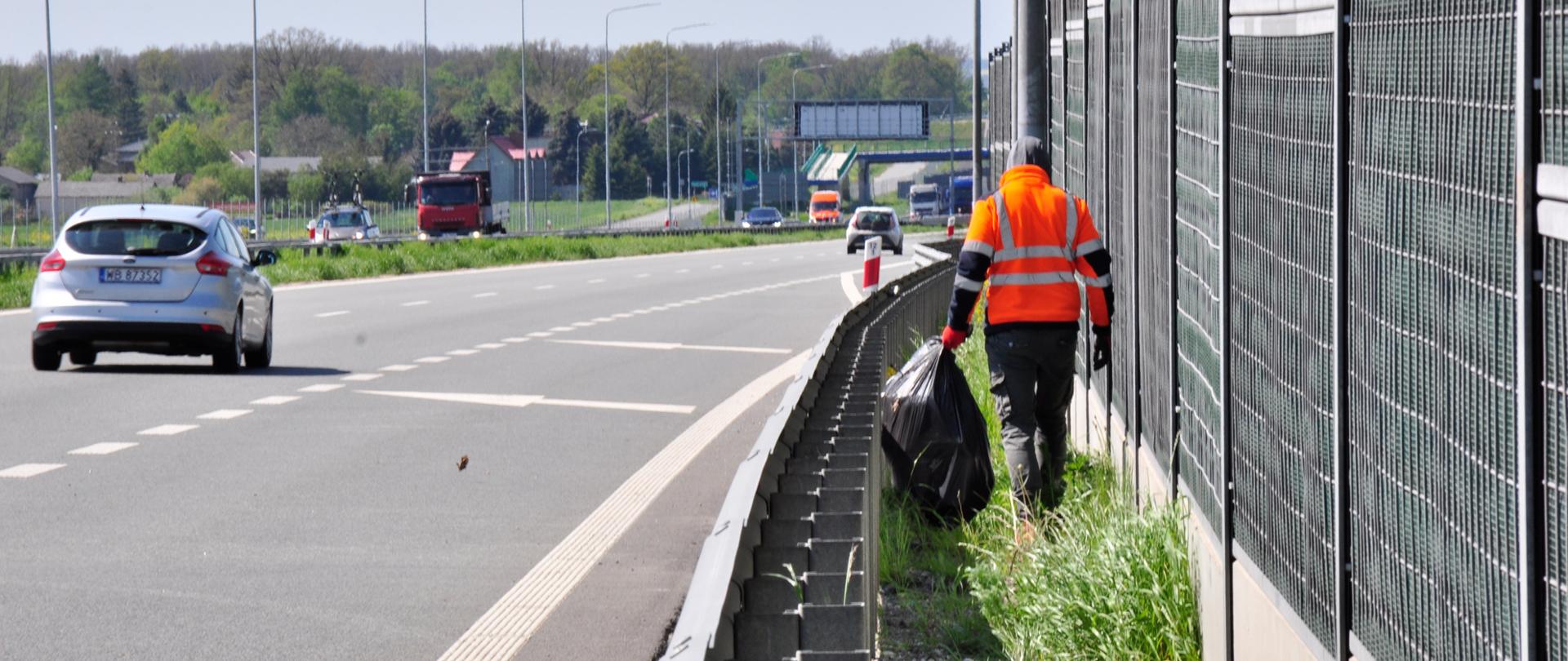 Zdjęcie przedstawia osobę ubraną w pomarańczową kamizelkę odblaskową, która porusza się wzdłuż pobocza autostrady, zbierając śmieci. Osoba ta trzyma duży czarny worek na śmieci i idzie obok metalowego ogrodzenia dźwiękochłonnego. Na drodze w tle widoczne są pojazdy, w tym srebrny samochód osobowy oraz czerwona ciężarówka. Po lewej stronie zdjęcia znajduje się pas zieleni oddzielający jezdnię od pobocza. W tle widoczne są drzewa oraz budynki, a dzień jest pogodny, z lekkim zachmurzeniem.