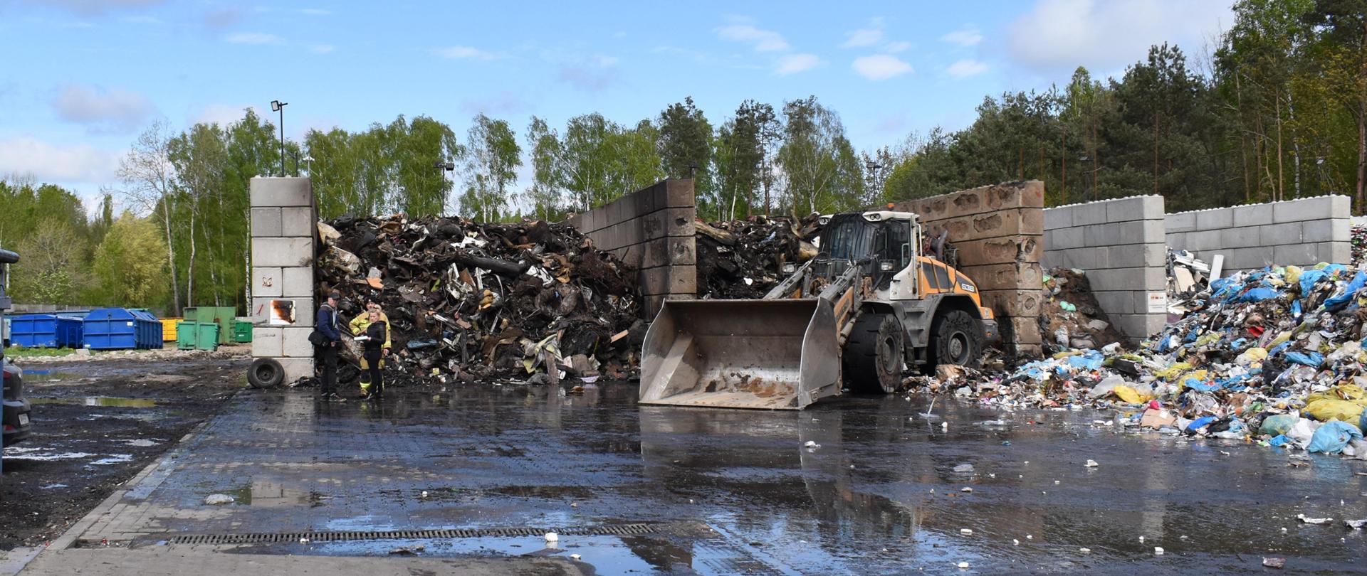 Spalone odpady wielkogabarytowe magazynowane w pryzmach o wysokości 4,5 m. Są one magazynowane w dwóch boksach. Przed nimi stoi spychacz. Po prawej stronie odpady, które nie uległy spaleniu.