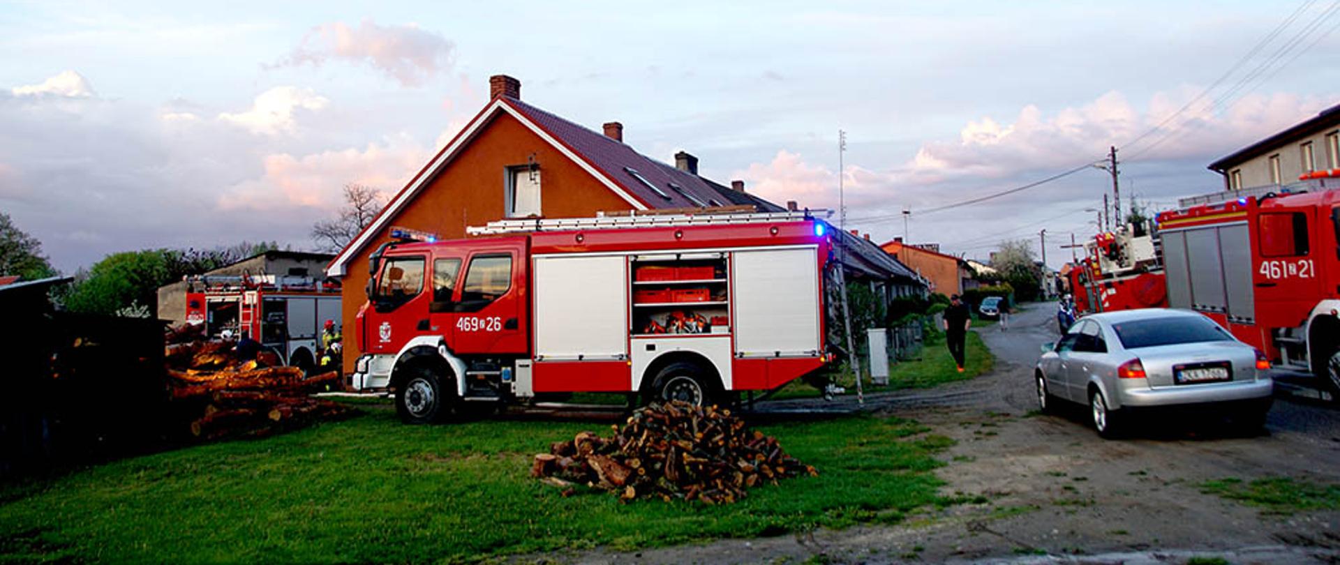Zdjęcie przedstawia zabudowania wielorodzinne, 4 wozy straży pożarnej, ulicę oraz jeden pojazd osobowy.