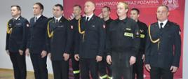 Uroczysta zbiórka z okazji NŚN. Zdjęcie przedstawia świetlicę Komendy Powiatowej PSP w Rawiczu. Na tle ścianki promocyjnej, w dwuszeregu stoi 10 strażaków. Część z nich ma nas obie mundur wyjściowy ze sznurem galowym, pozostali - ubrania koszarowe. 