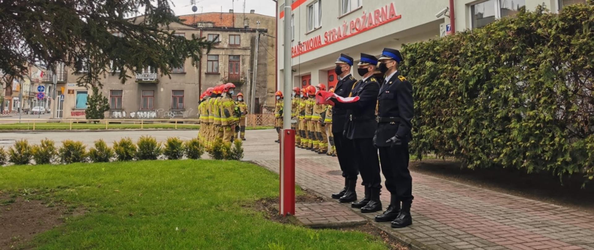 Poczet flagowy w mundurach wyjściowych ze złożoną flagą przed masztem flagowym.