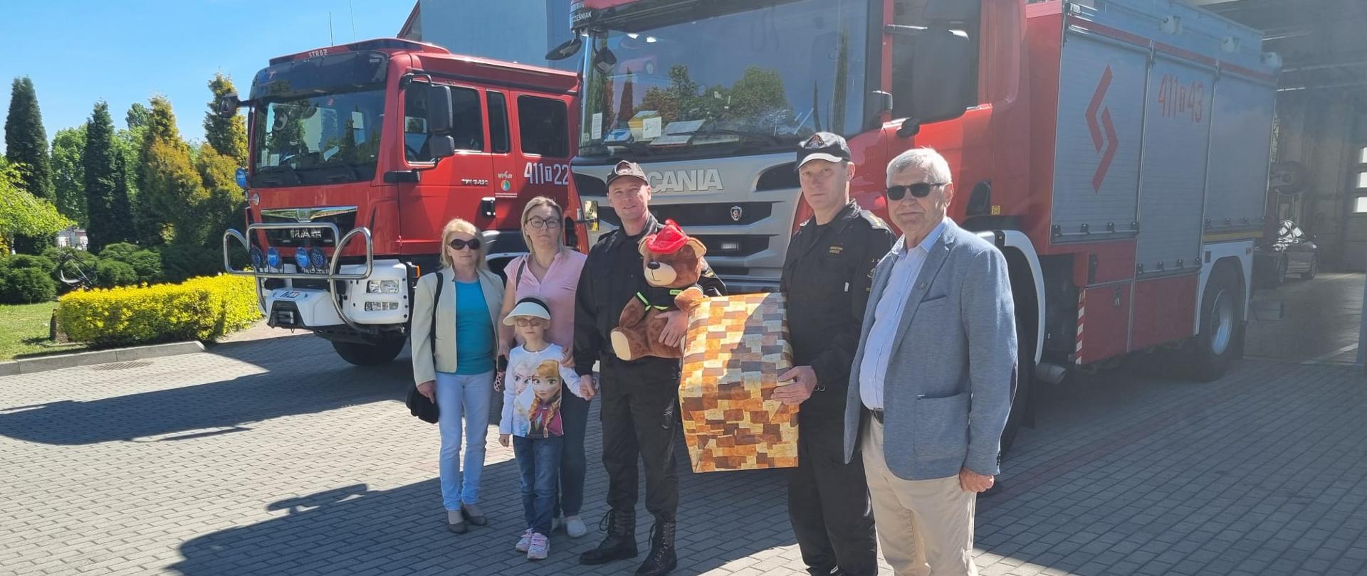 Na zdjęciu obok dwóch pojazdów ratowniczo-gaśniczych ustawieni są; komendant powiatowy PSP w Jędrzejowie, dziewczynka wraz z opiekunami, dowódca Jednostki Ratowniczo-Gaśniczej w Jędrzejowie oraz pracownik Polskiego Związku Niewidomych. Strażacy trzymają prezent oraz pluszowego misia.