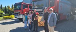 Na zdjęciu obok dwóch pojazdów ratowniczo-gaśniczych ustawieni są; komendant powiatowy PSP w Jędrzejowie, dziewczynka wraz z opiekunami, dowódca Jednostki Ratowniczo-Gaśniczej w Jędrzejowie oraz pracownik Polskiego Związku Niewidomych. Strażacy trzymają prezent oraz pluszowego misia.
