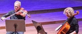 Dwóch mężczyzn siedzi na krzesłach na estradzie grając na altówce i wiolonczeli. Przed nimi stoją pulpity.