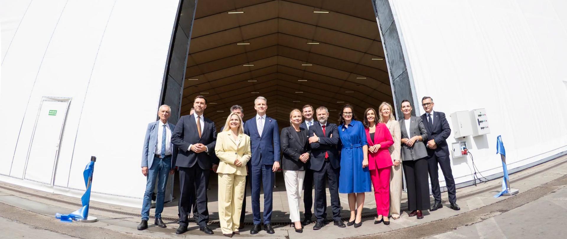 Grupa osób stoi i uśmiecha się przed wejściem do wielkiej, białej hali namiotowej. W tle widać doświetlone wnętrze obiektu z widoczną konstrukcją dachu.