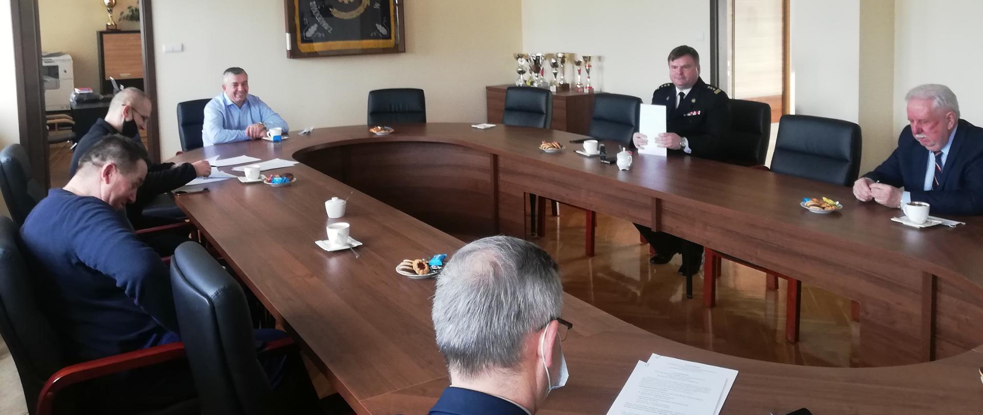 Przy stole na sali wykładowej siedzą mężczyźni. Na drewnianym stole poczęstunek oraz dokumenty.