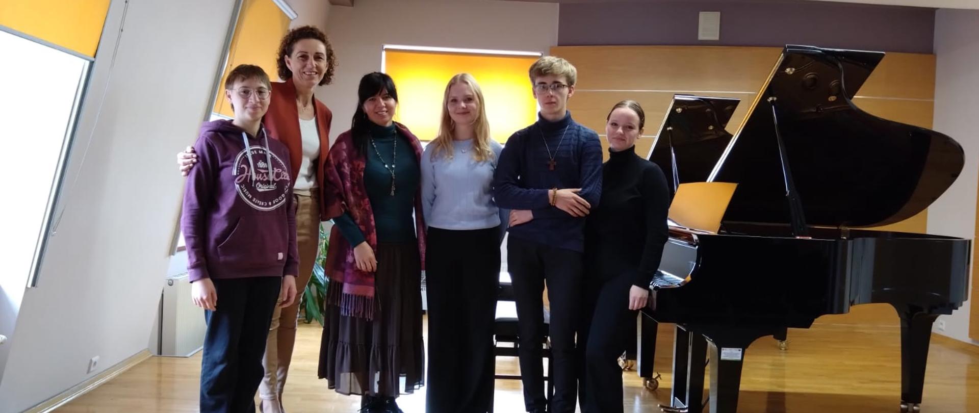 Zdjęcie grupowe uczestników warsztatów pianistycznych z nauczycielem oraz prowadzącej warsztaty, którzy stoją w jednym rzędzie. Za nimi dwa fortepiany oraz ławy fortepianowe oraz okno zasłonięte roletami. Z boku dwa okna również przysłonięte roletami oraz kaloryfer
