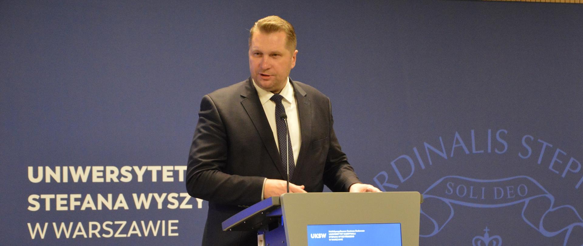 Minister Czarnek stoi przy mównicy i mówi do mikrofonu, za nim na fioletowej ścianie napis Uniwersytet Stefana Wyszyńskiego w Warszawie.
