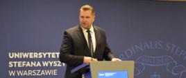 Minister Czarnek stoi przy mównicy i mówi do mikrofonu, za nim na fioletowej ścianie napis Uniwersytet Stefana Wyszyńskiego w Warszawie.