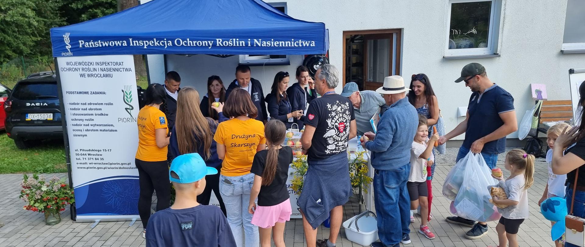 Przed budynkiem świetlicy wiejskiej odbywa się plenerowe wydarzenie. Przy namiocie z napisem „Państwowa Inspekcja Ochrony Roślin i Nasiennictwa” zgromadzili się dorośli i dzieci. Pracownicy stoiska rozmawiają z uczestnikami i przekazują materiały. Wokół stoiska stoją osoby w różnym wieku, w tym dzieci; część uczestników trzyma torby i materiały promocyjne. 
