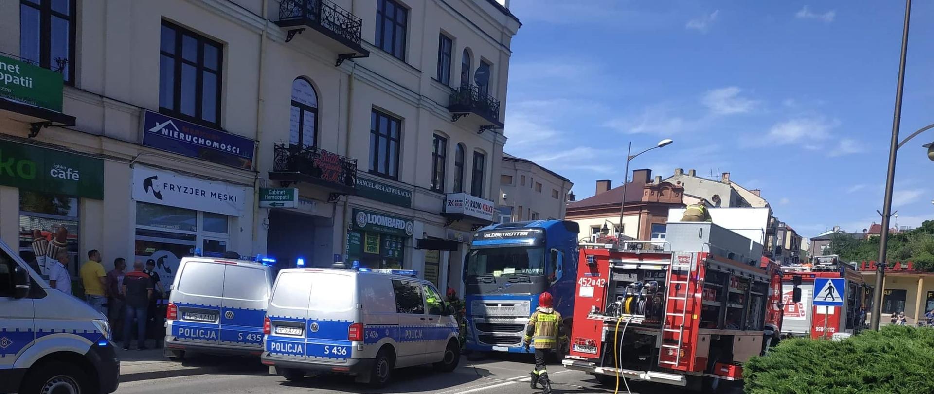 Na pierwszym planie 3 radiowozy policyjne, 2 samochody strażackie, zablokowana ulica. Za pojazdami służb ciężarówka pod którą znajduje się uwiezione ciało kobiety. W tle kamienice miejskie i przechodnie. 