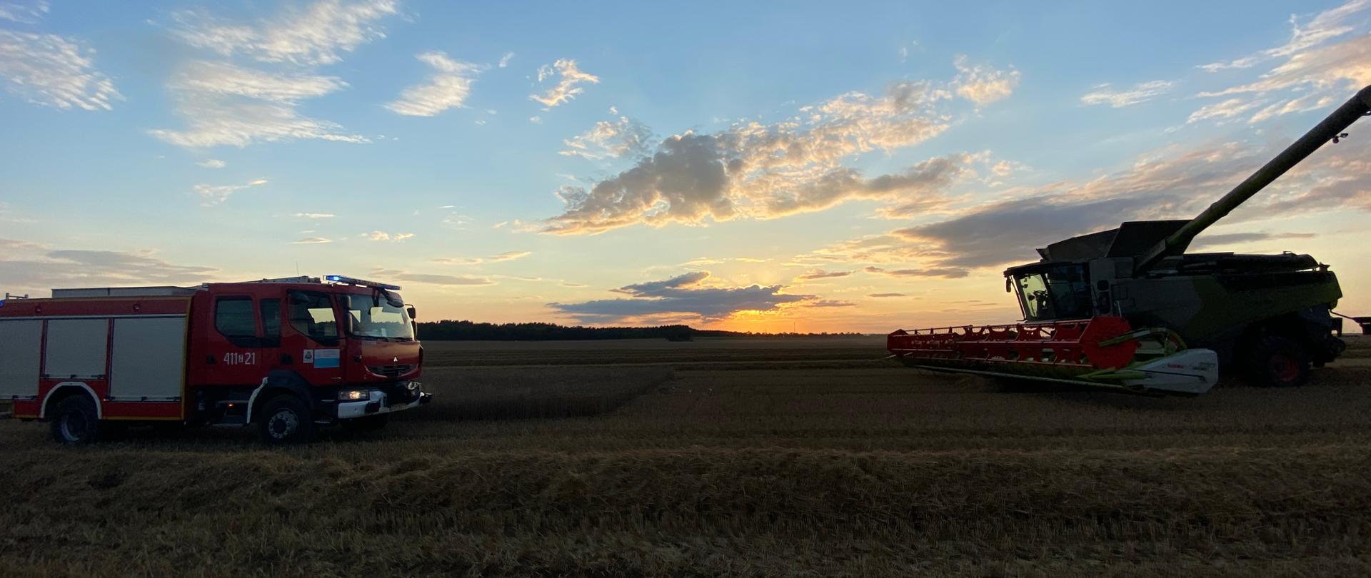 Na fotografii po lewej stronie znajduje się wóz strażacki, który stoi na polu - pole jest ścierniskiem. Wóz jest czerwony i ma włączone niebieskie sygnały świetlne. Po prawej stronie stoi biało-zielony kombajn, który ma opuszczony heder. Zdjęcie wykonano przy zachodzie słońca.